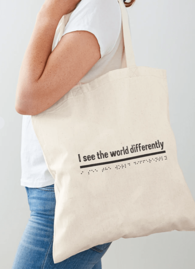 A woman with a white t shirt and blue jeans, stands to the side and has a beige tote bag over her shoulder. The words on the bag are I see the world differently