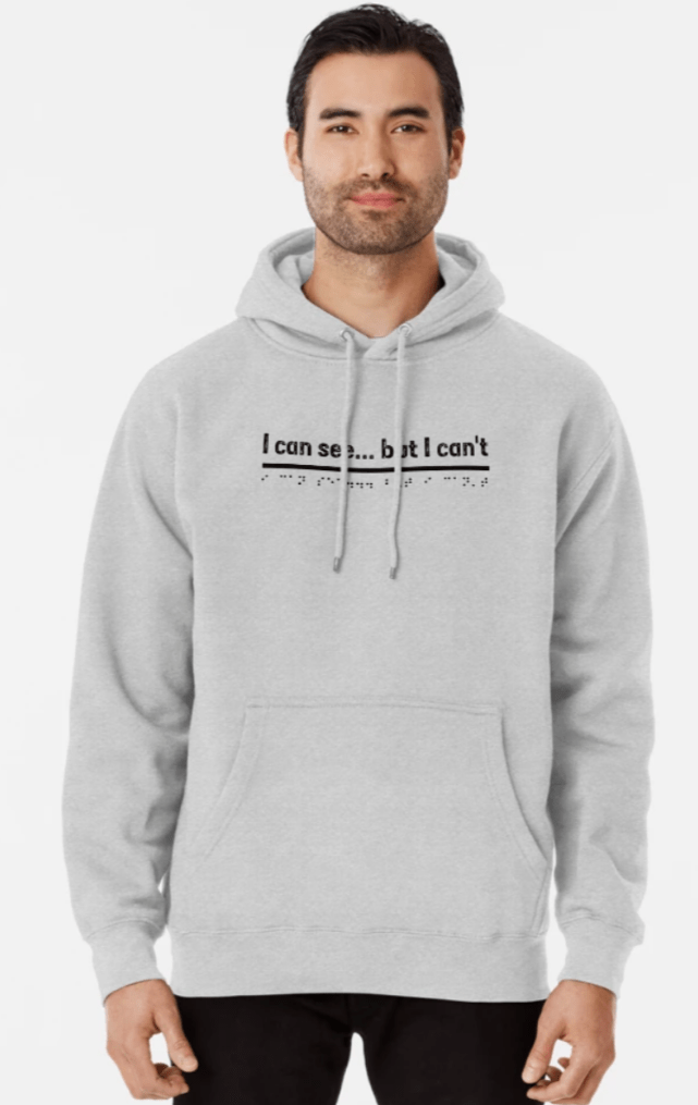A man stands with his arms by his side. He has brown hair and a bit of stubble on his face. He wears black trousers and a grey hoodie jumper. The jumper has the words. I can see but I can't in black with the equivalent Braille font underneath.