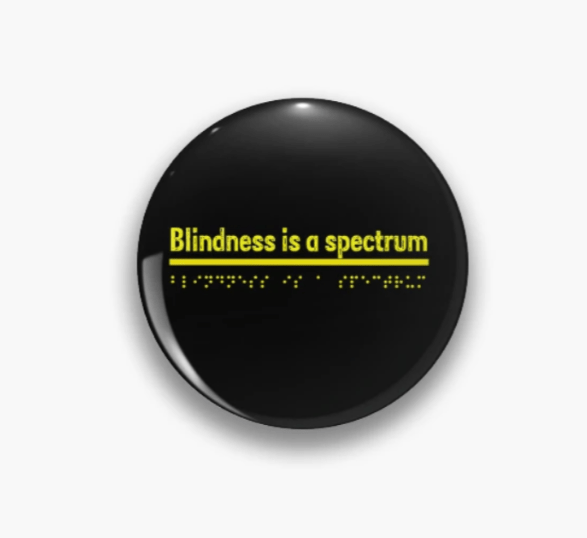 A black badge pin button on a white background. The words on the badge read Blindness is a spectrum in yellow with same equivalent Braille font underneath