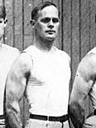 Black and white photograph of George standing slightly diagonally, hands behind his back. He wears a tight white vest top and light coloured trousers with a belt. He has deep set eyes and very short light slicked hair.