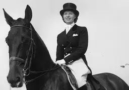 Black and white photograph of a short curly style dark haired woman sitting on a dark coloured horse. She’s smiling and wears a smart cropped jacket, top hat and white jodhpurs.