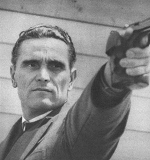 Black and 2hote photograph of Karoly Takacs . Arm outstretched holding a gun. A dark haired man with slicked back hair. A serious look on his face concentrating. Wearing a jumper and blazer.
