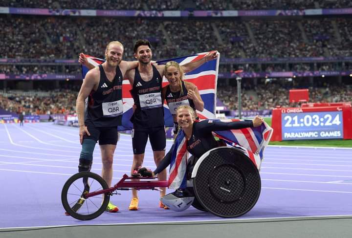 with the stadium in the background and a union jack behind the 4 Paralympians in the team. 2 men, Jonnie and Zac, with arms around each other smiling. Blonde woman, Ali, slightly crouching down smiling. Blonde woman Sammi sat in her racing wheelchair smiling whilst holding the flag