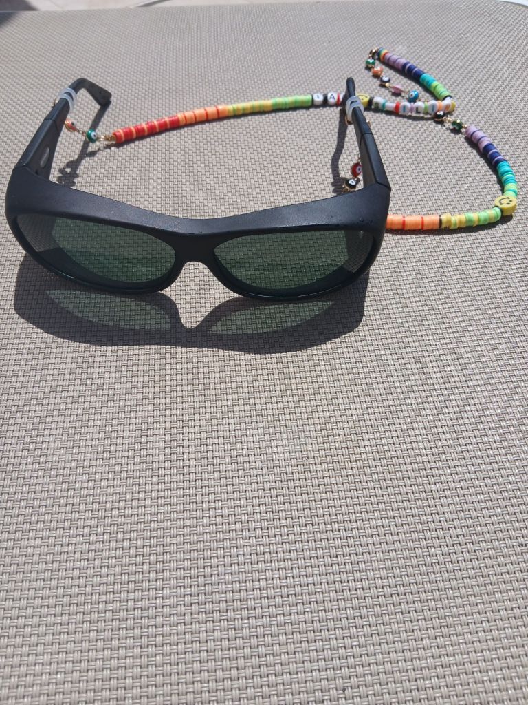 A pair of black Cocoon sunglasses placed on a grey mesh sunlounger from the top view. They have a multicoloured string of beads attached to the ends of them to wear around the neck.