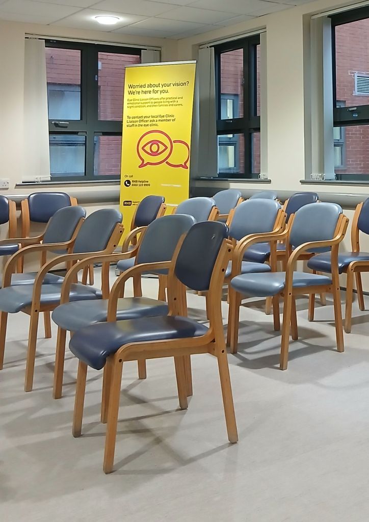 A hospital waiting room with blue and light wood chairs in the foreground. Windows are in the background with a large yellow poster with text that reads. Worried about your vision? We're here for you.
