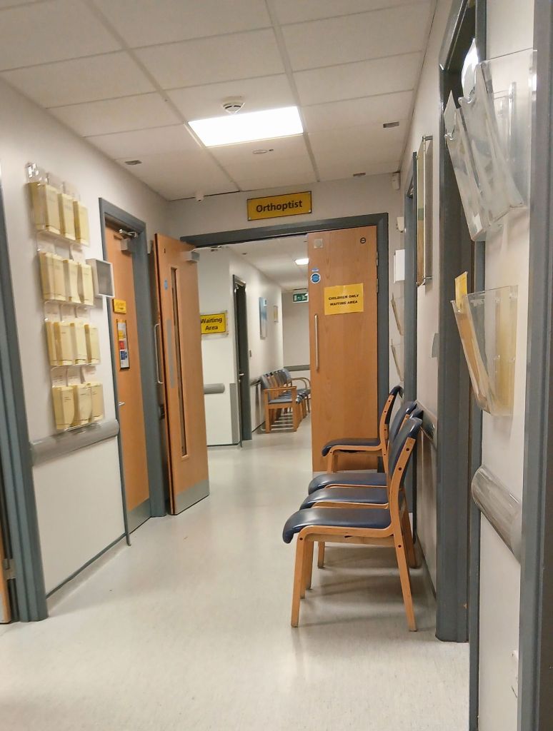 A hospital corridor with chairs down the right hand side. Above double doors is a sign that reads Orthoptist. 