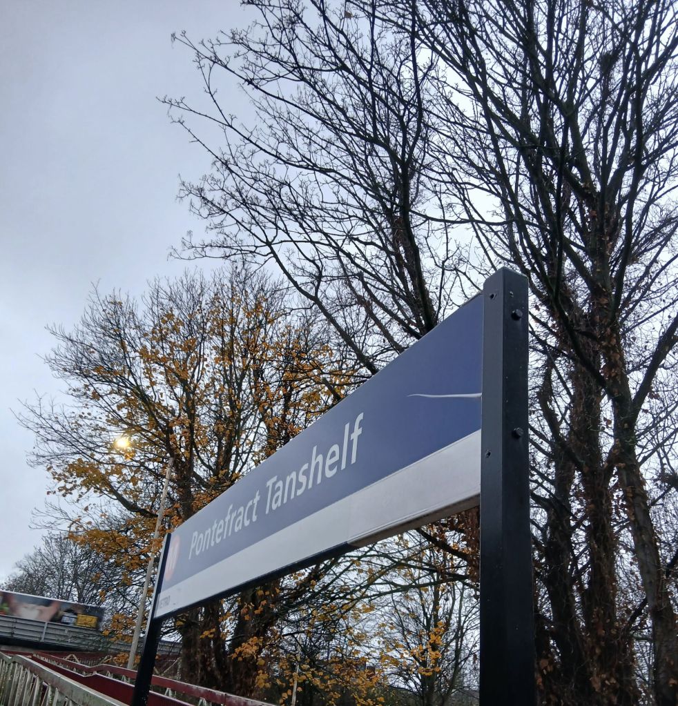 Close up view of the train station sign. It reads Pontefract Tanshelf. Behind the sign are trees against the sky. A yellow blob looks to be behind the trees. It's a light with a light coloured pole.