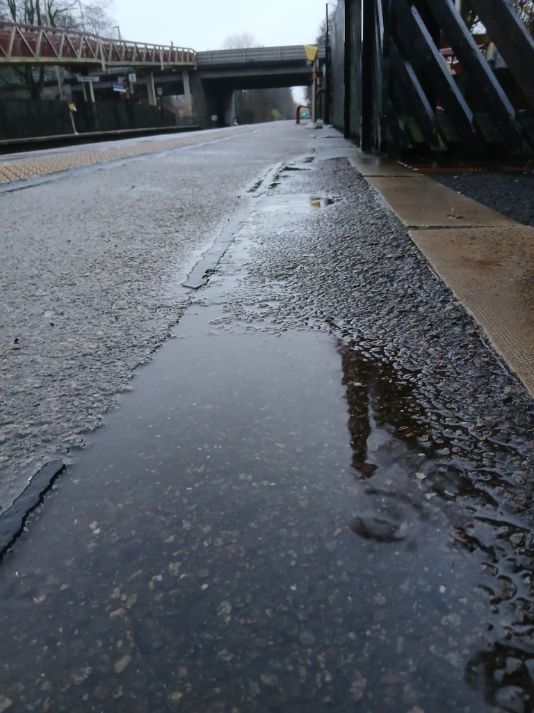 PD 3 A low down shot of the station with the tracks and bridge in the distance. A large puddle is in the very near foreground. 