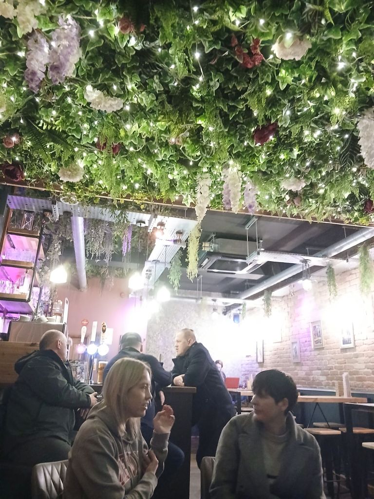 Two young women sat together facing each other at the foreground. One with short blonde hair and one with short dark hair chat infront of three people in the background sat at the bar. At the top of the picture there is green foliage and flowers on the ceiling filled with fairy lights.