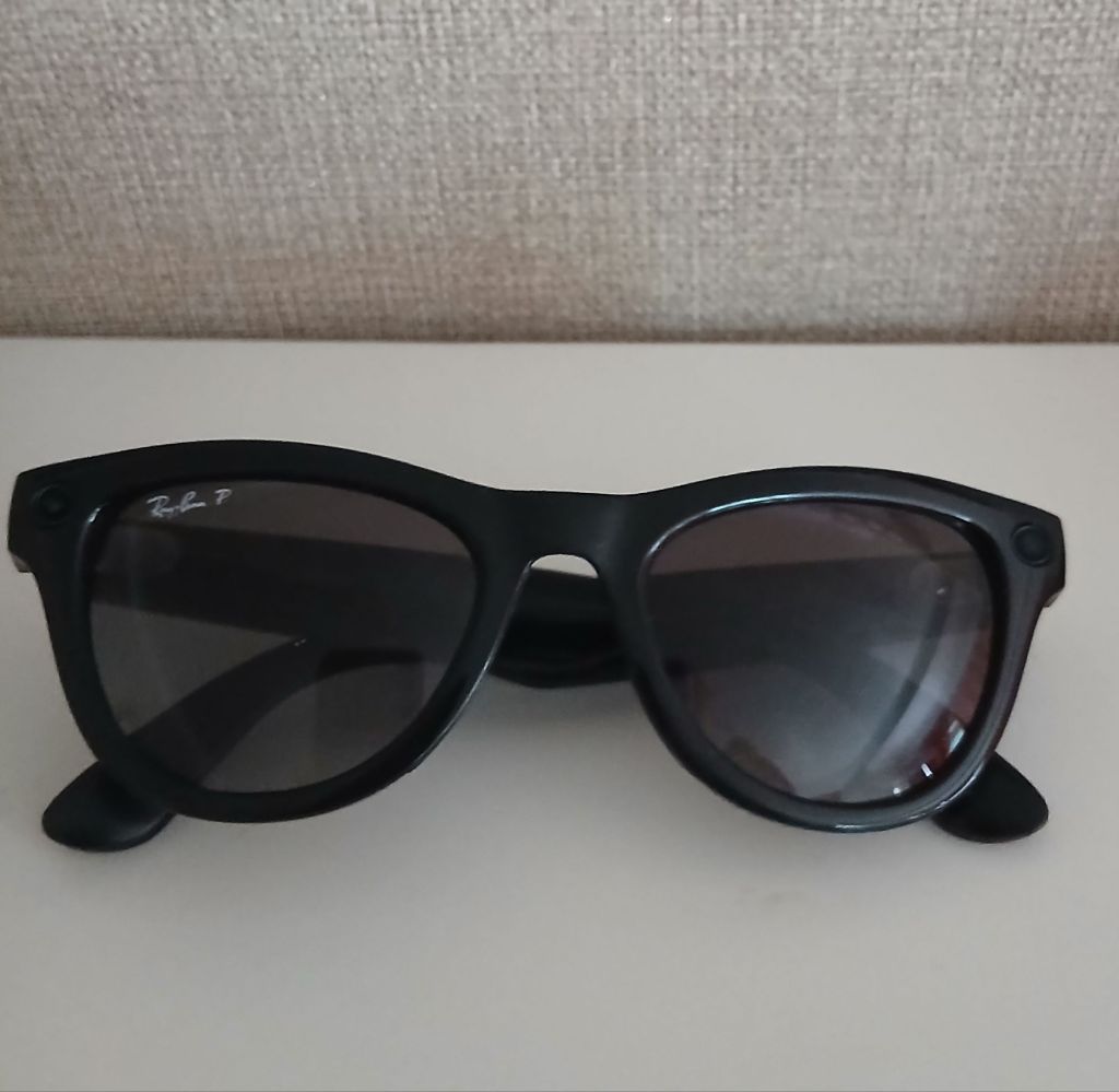 A pair of black Ray-Ban Meta Skylar sunglasses sit on a white surface. Behind in the background is a beige coloured fabric wallpaper. The glasses are folded up and are a cats eye winged shape. 