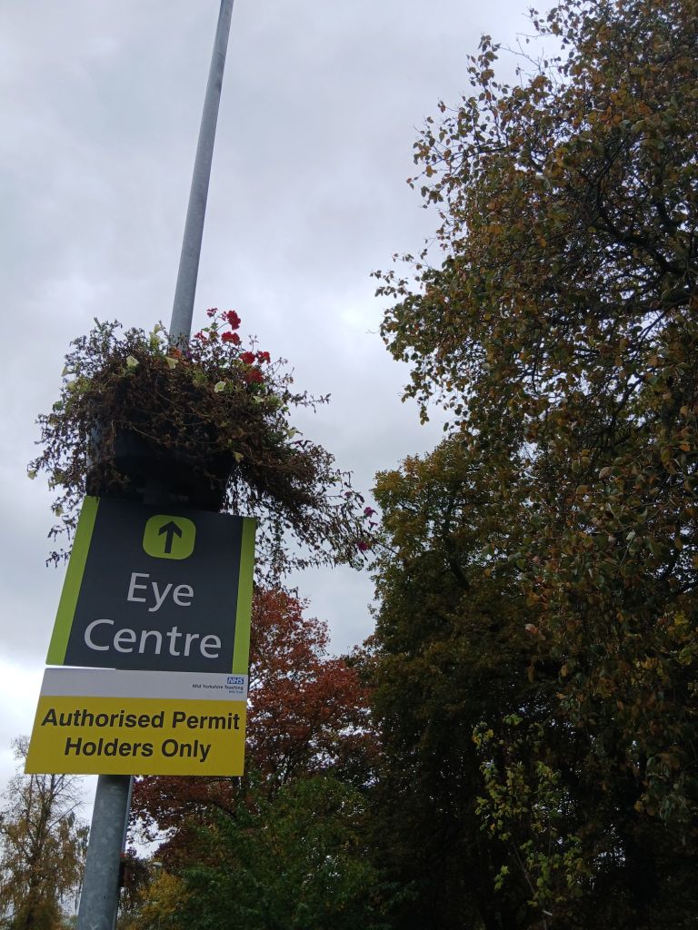 Standard photo of the Eye Centre sign in green and grey. Autumn trees behind in green yellow and orangey red